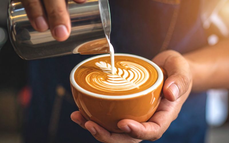 A barista's hands pouring perfectly steamed milk from a pitcher into a cup of espresso, creating a beautiful latte art heart.