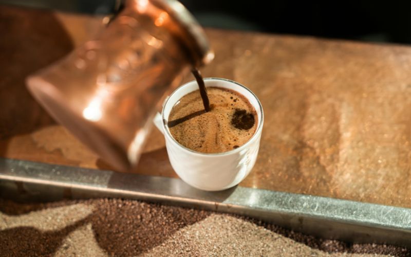 A traditional copper Turkish coffee pot, known as a cezve, pouring rich, foamy coffee into an ornate cup.